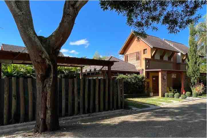 The exterior of a welcoming villa is showcased, featuring a blend of brick and wooden elements. A lush tree provides shade to the property, while a wooden fence delineates the space. The clear blue sky complements the warm tones of the building.