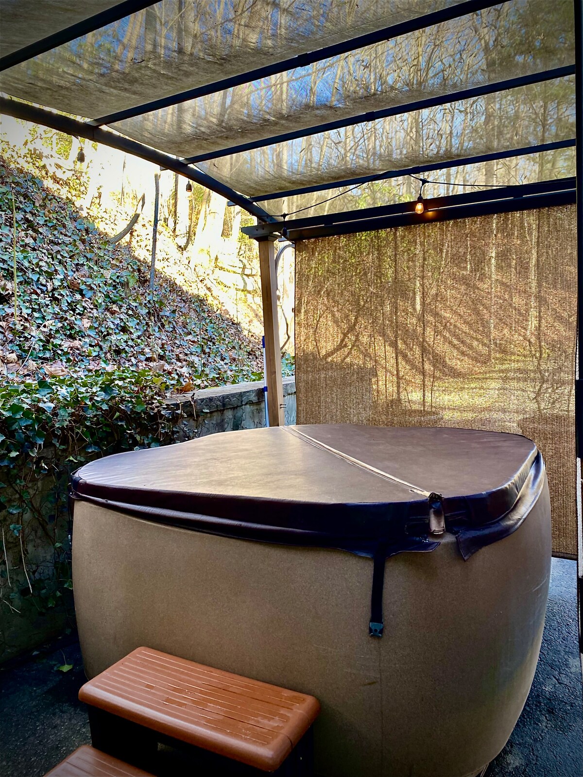 A private hot tub is seen under a canopy, surrounded by natural greenery and a wooden deck. The walls are partially draped with a shade cloth, providing privacy. Soft lighting fixtures hang above, enhancing the relaxing atmosphere of this outdoor retreat.