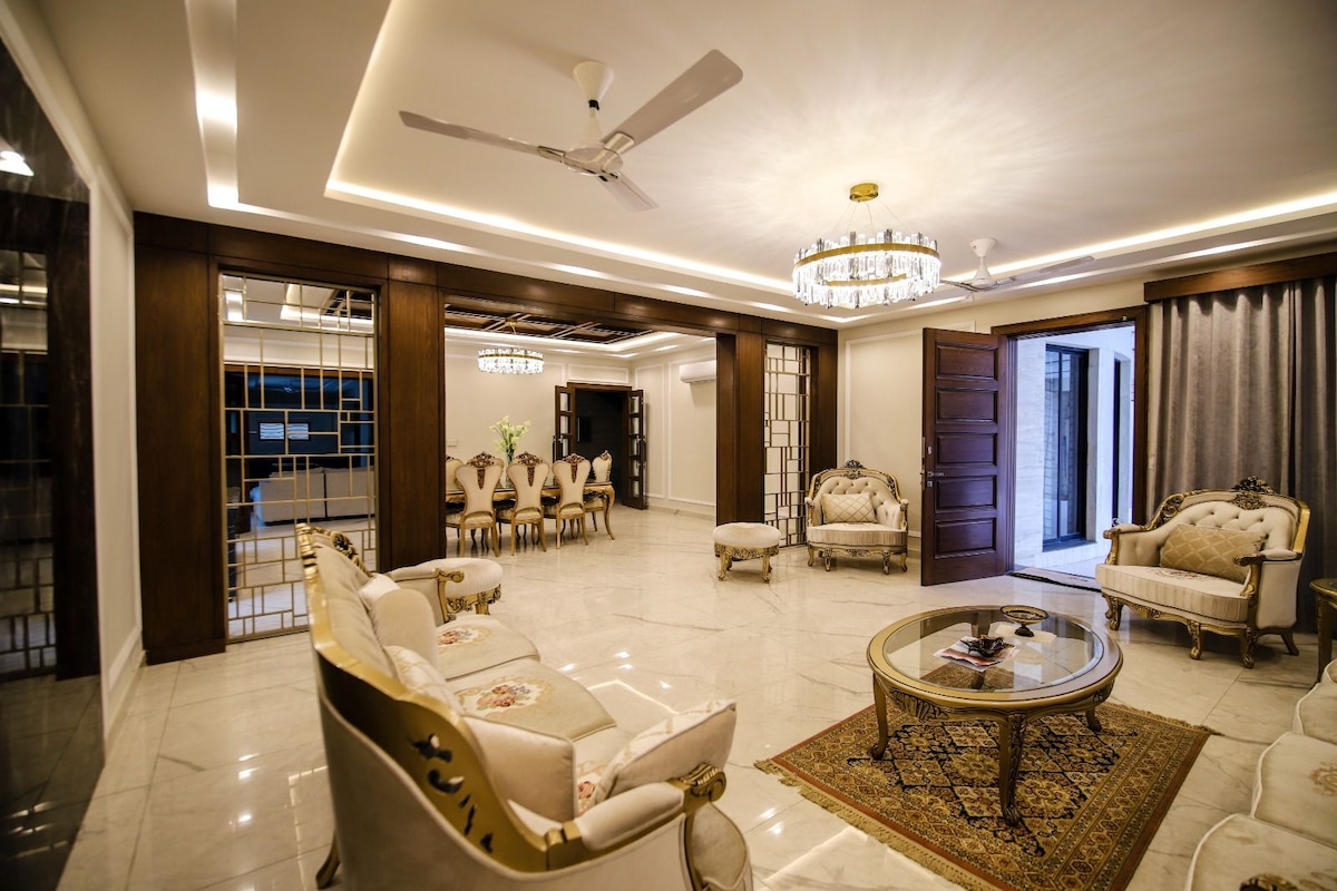 A spacious and elegantly furnished living area features a circular coffee table at its center. Surrounding seating includes light-toned sofas and armchairs with decorative accents. Soft lighting is provided by a crystal chandelier, while large glass doors allow natural light to enter from the outside.