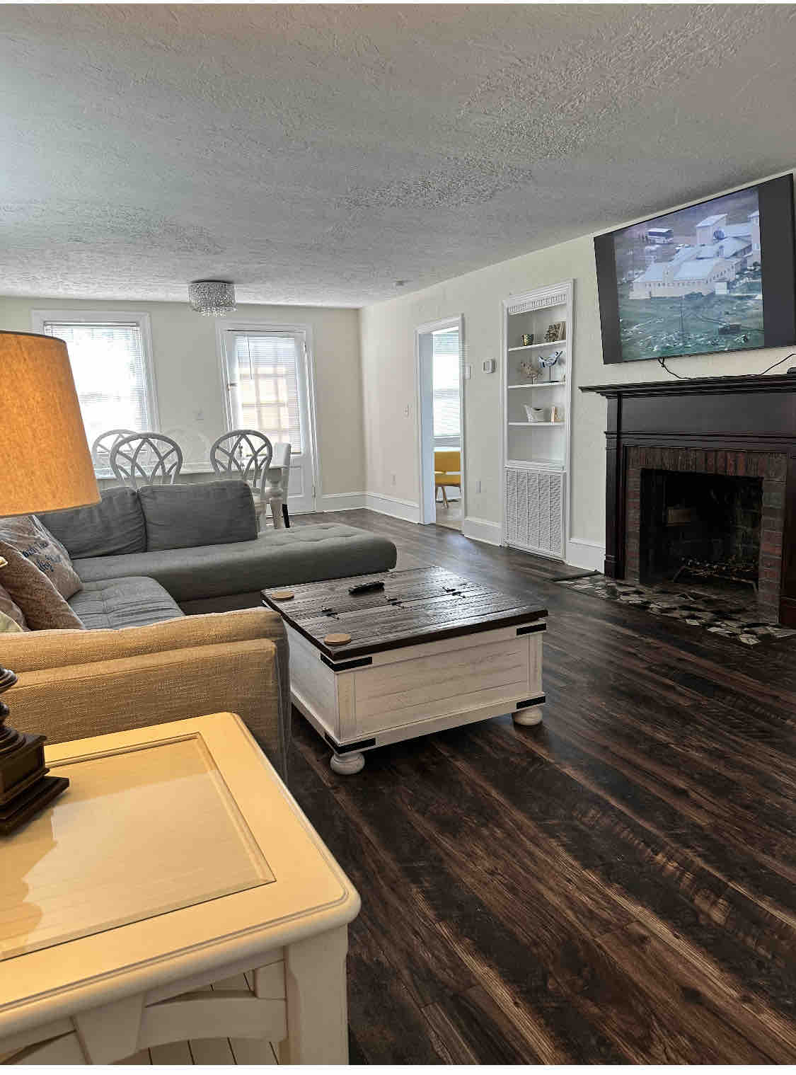A living area features a comfortable sectional sofa complemented by a decorative lamp and a white storage table. A television is mounted above a dark fireplace, while natural light enters through large windows. The space is designed for relaxation and social gatherings.
