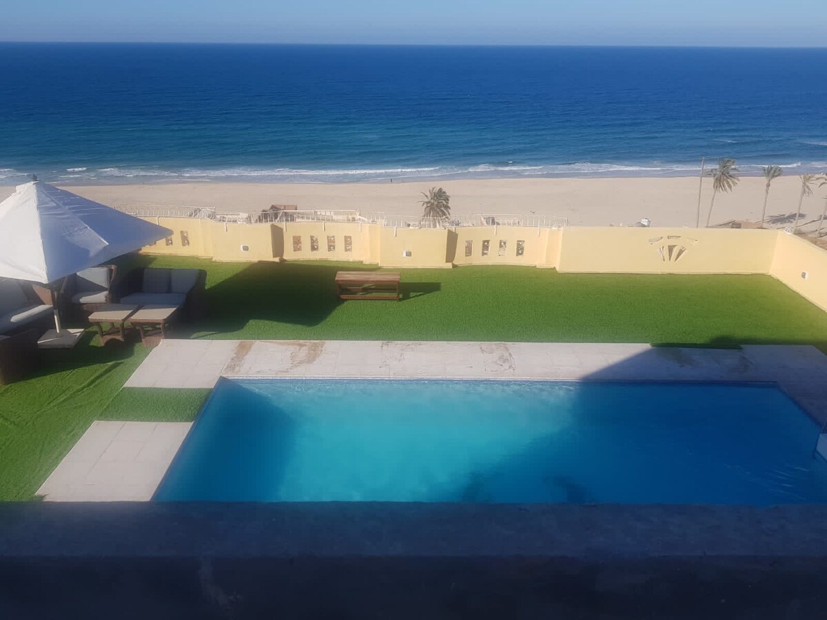 An expansive view showcases a private pool surrounded by a manicured lawn, leading to a sandy beach and open sea. Lounge chairs and patio furniture are arranged for relaxation under a shaded structure, enhancing the outdoor living space.