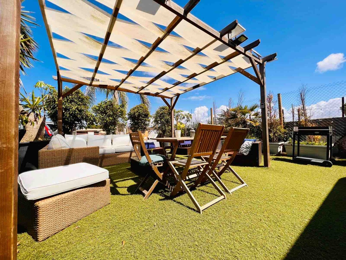 A covered outdoor seating area is depicted, featuring a mix of lounge furniture and dining tables. Green artificial turf enhances the space, while various plants surround it. A clear blue sky is visible above, providing a bright and airy atmosphere.