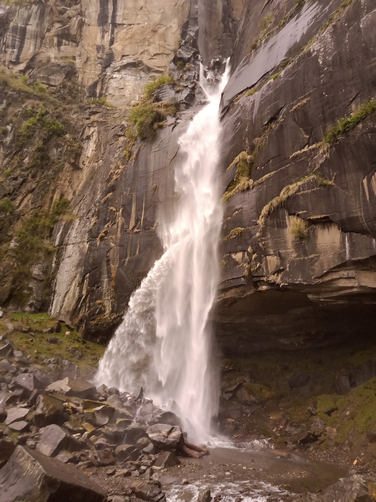 A cascading waterfall tumbles down rocky cliffs, surrounded by textured stone formations. The water flows rapidly, creating a mist at the base, where smooth stones are scattered on the ground. Lush greenery is present along the rocky edges, maintaining a natural aesthetic.