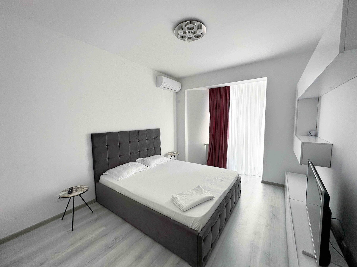 A spacious bedroom features a large, upholstered bed complemented by a side table. A flat-screen TV is mounted on the wall opposite the bed. Natural light enters through a window, which is partially covered by a curtain, and a sleek air conditioning unit is installed for comfort.