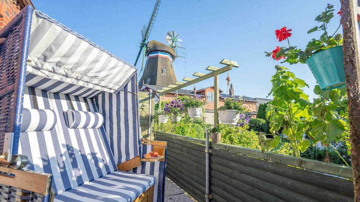 A striped beach chair sits in a well-kept garden, surrounded by potted plants and herbs. A windmill stands in the background under a clear blue sky. The cozy seating area is shaded, creating a relaxing spot to enjoy the tranquil surroundings.
