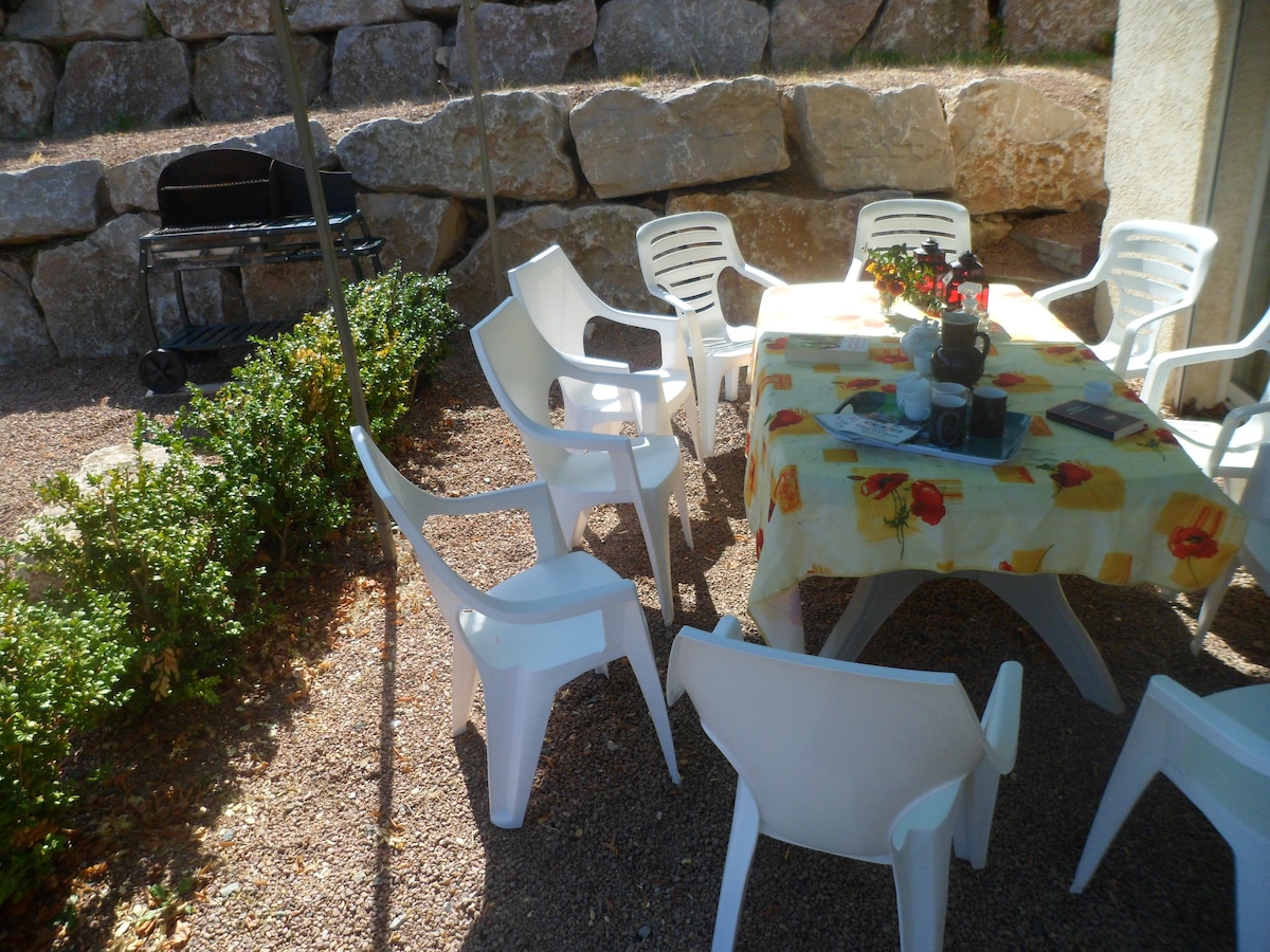 An outdoor dining area is shown, featuring a table covered with a floral tablecloth surrounded by several white chairs. A barbecue grill is positioned nearby, with natural stone walls providing a rustic backdrop. Green shrubbery edges the space, enhancing the serene environment.