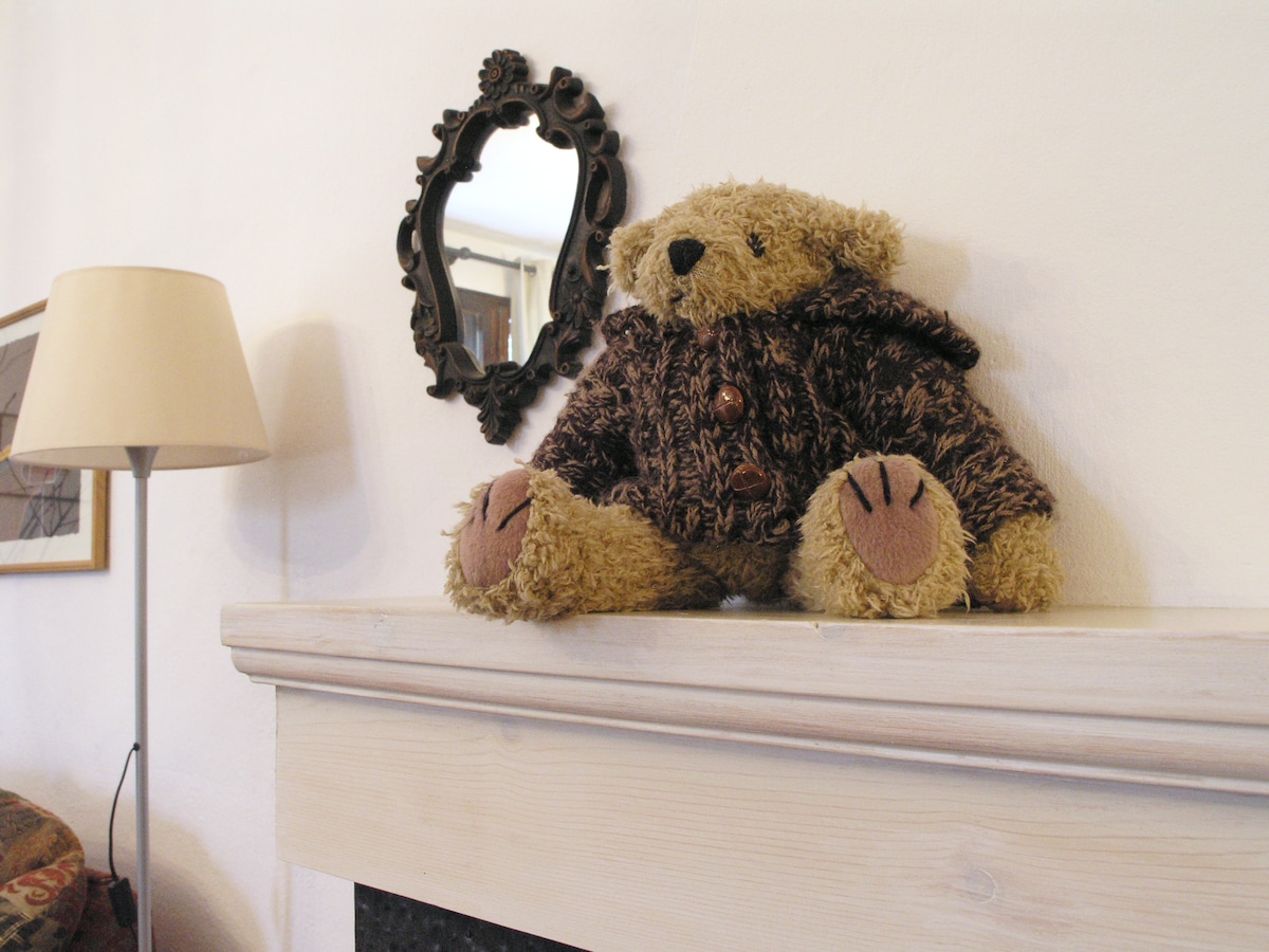 A soft teddy bear dressed in a dark coat is positioned on a wooden mantel. The bear is framed by a small, ornate mirror hanging on the wall behind it. A minimalist lamp stands to the left, adding to the room's decor.