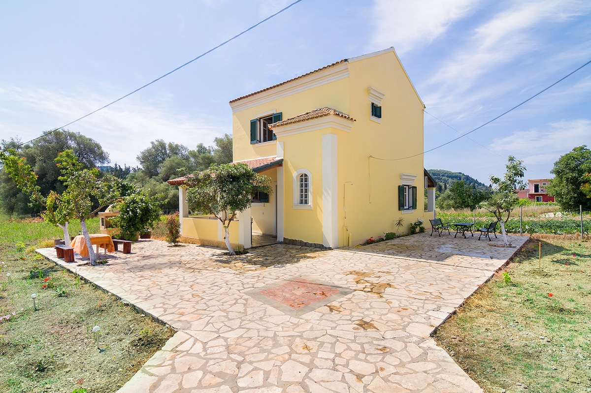 A two-story yellow villa is set within a green landscape, featuring a spacious stone patio surrounded by trees. Chairs and a small table are arranged for outdoor gatherings. The villa is designed with multiple windows, providing natural light and a view of the serene surroundings.