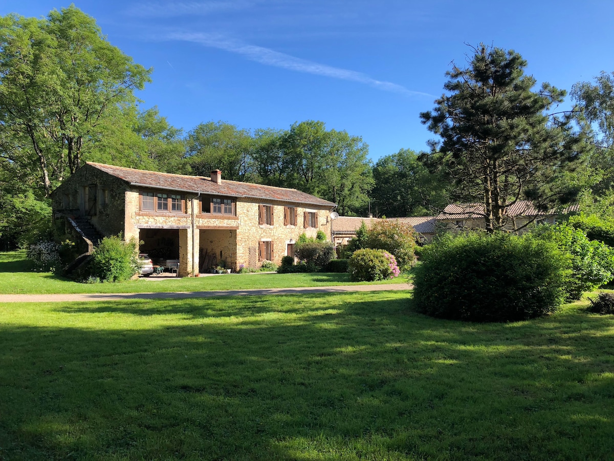 A charming stone farmhouse is set amidst lush greenery, featuring a two-story facade with wooden shutters. The landscape is characterized by vibrant shrubs and trees, complementing the tranquil setting. A well-maintained lawn extends in front, inviting relaxation in the natural surroundings.