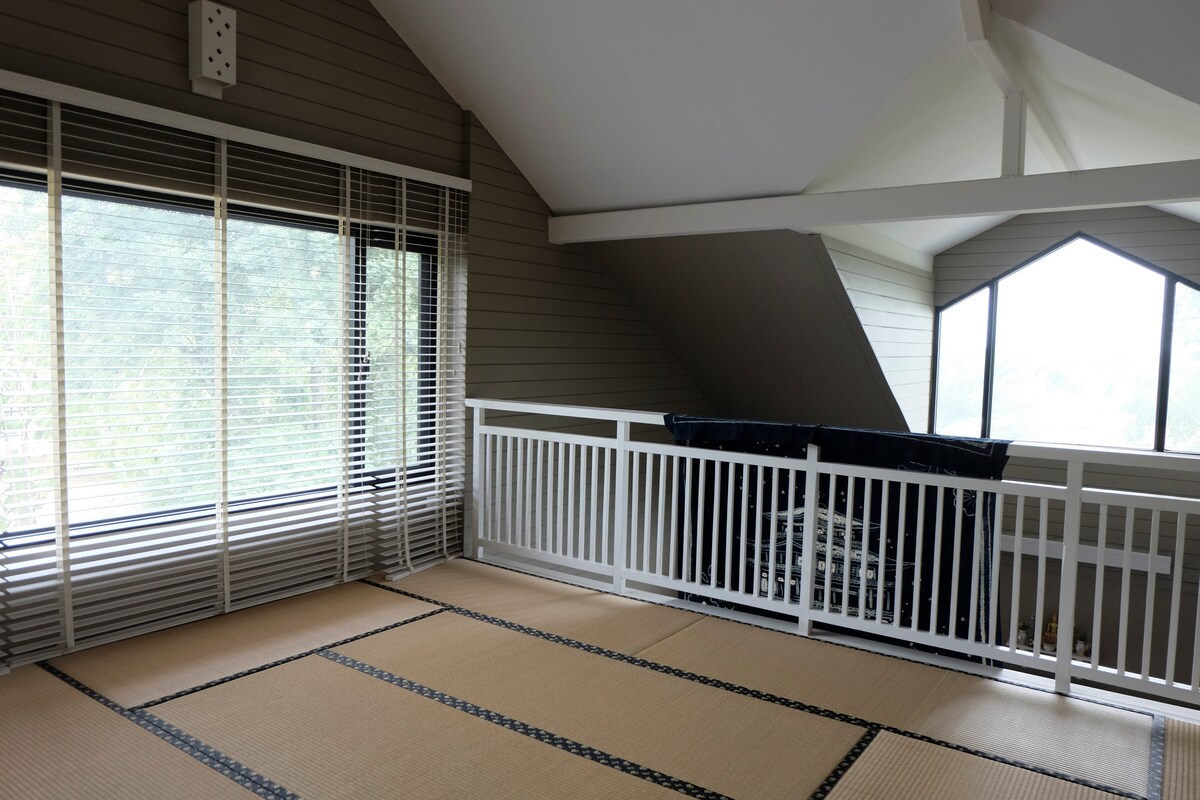 A serene mezzanine level features natural light streaming through large windows adorned with blinds. The floor is covered in traditional tatami mats, offering a calm and minimalistic atmosphere. A wooden railing provides safety while allowing views of the surrounding landscape.