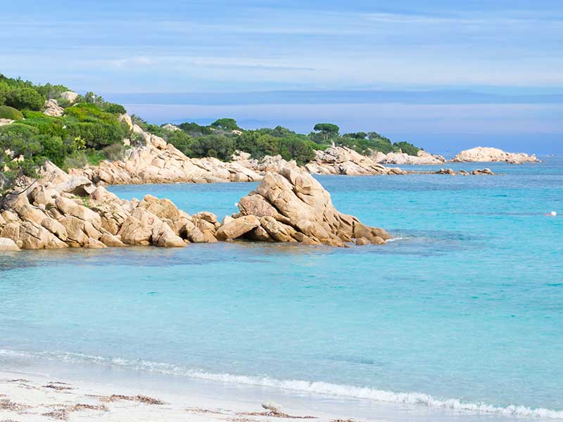 A serene beach landscape is showcased, featuring gentle waves lapping against a sandy shore. Smooth rocks emerge from the turquoise water, and lush green hills line the background, contributing to the tranquil atmosphere.