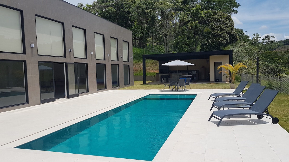 A modern outdoor area features a rectangular swimming pool surrounded by a spacious patio. Comfortable lounge chairs are positioned nearby. An outdoor dining area with a shaded table and chairs is visible under a canopy, bordered by manicured landscaping and trees.