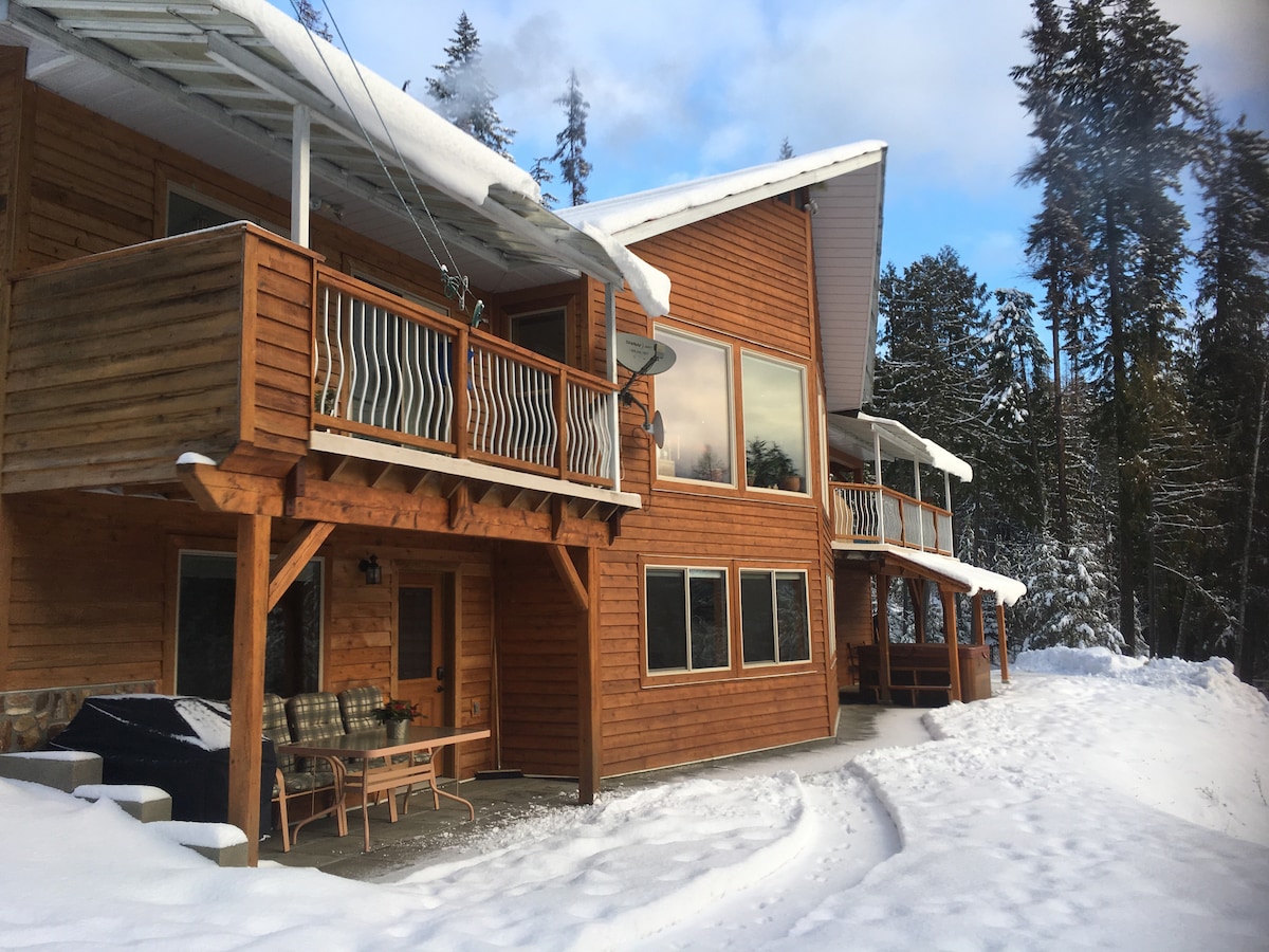 The exterior of the property is shown, featuring a combination of wood siding and multiple balconies. Snow covers the ground, while tall trees surround the area. A patio set is visible, and a pathway leads toward the entrance, enhancing the sense of tranquility.
