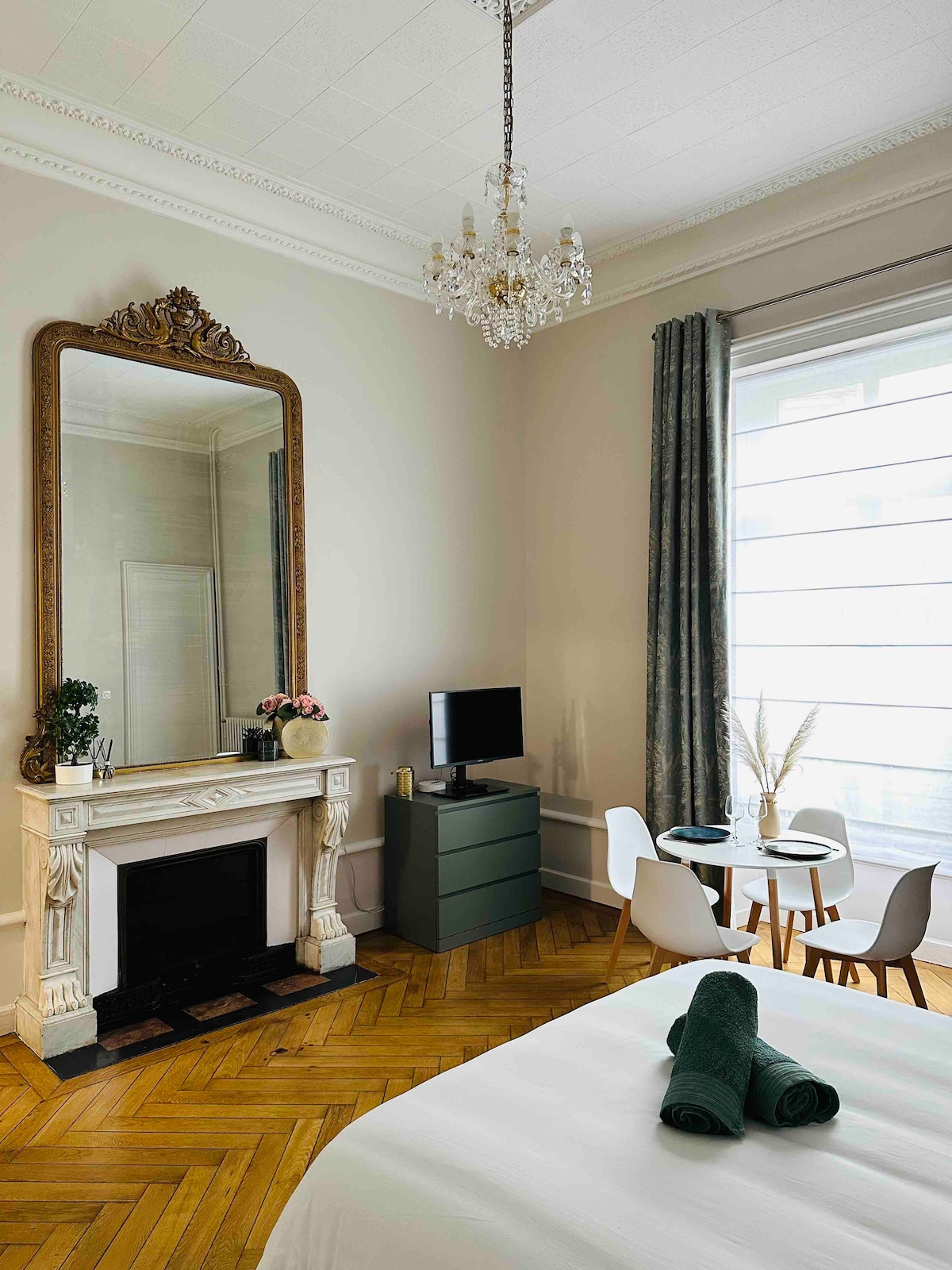 A spacious room features an elegant mirror framed in gold above a marble fireplace. A small dining table with white chairs sits next to a modern television and a dark green cabinet. Natural light filters through sheer curtains, highlighting the patterned wooden floor.