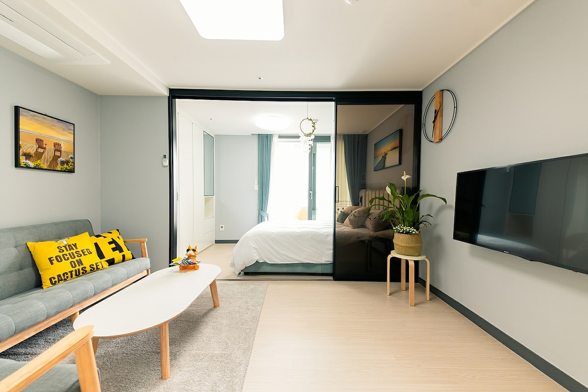 A modern living area is featured, with a comfortable gray sofa positioned beside a minimalistic wooden coffee table. A large flat-screen TV is mounted on the wall opposite. Natural light enters through glass sliding doors, leading to the sleeping area, creating a bright and open space.