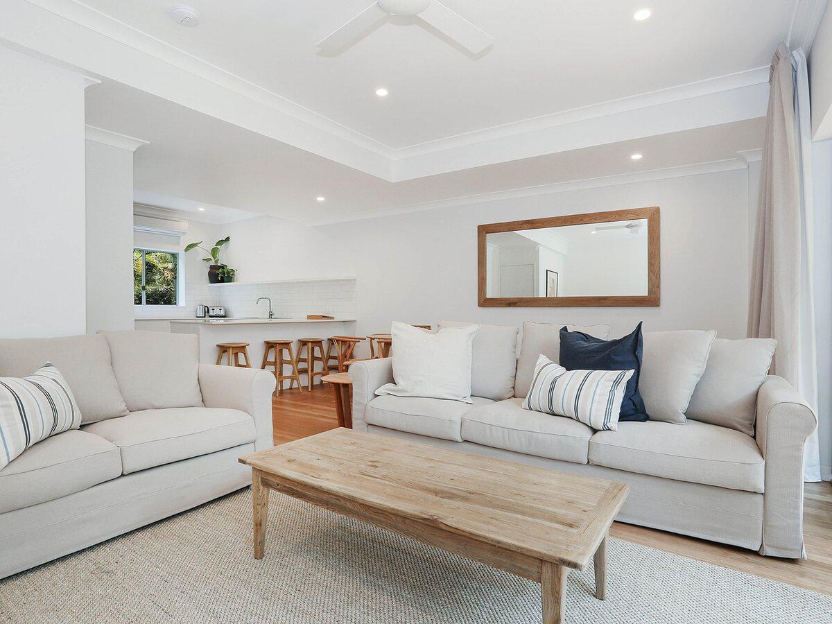 A spacious living area features two light-colored sofas adorned with striped pillows. A wooden coffee table is positioned in the center, reflecting natural tones. In the background, a modern kitchen is visible with light-colored cabinetry and wooden stools, complemented by bright overhead lighting.