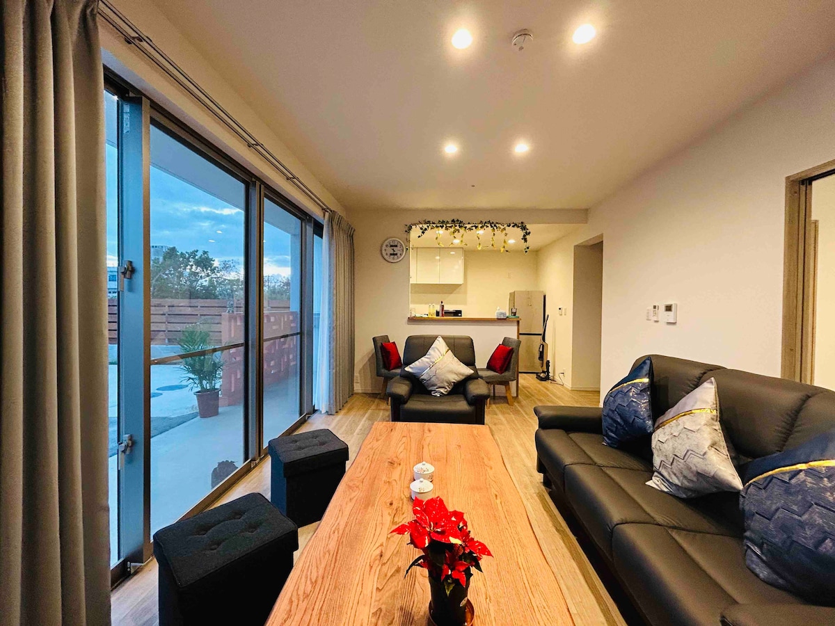 A modern living area features a wooden coffee table at the center surrounded by black leather sofas. A bright kitchen area is visible in the background, with a view of the outdoor space through large glass doors allowing natural light to fill the room.