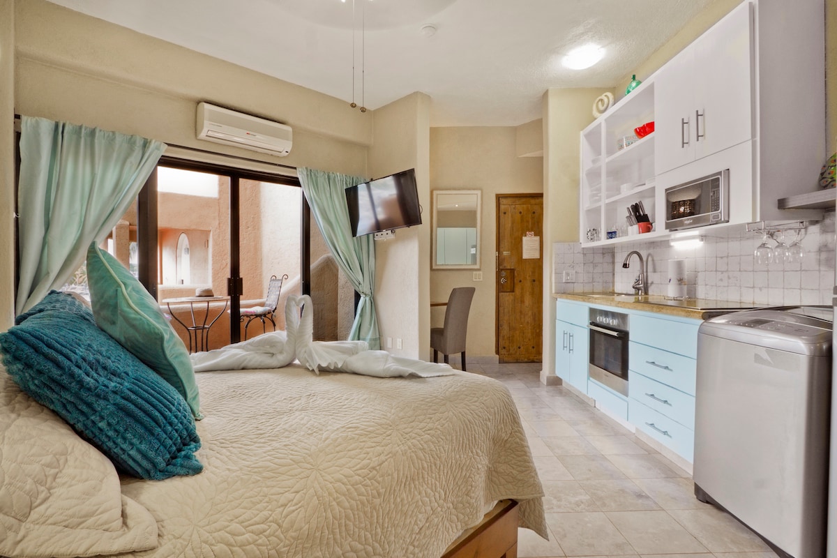 A cozy studio apartment features a spacious bed with soft pillows and a textured quilt. Light blue cabinets complement a functional kitchenette that includes modern appliances. Natural light streams in through the large sliding doors, leading to a small outdoor terrace.