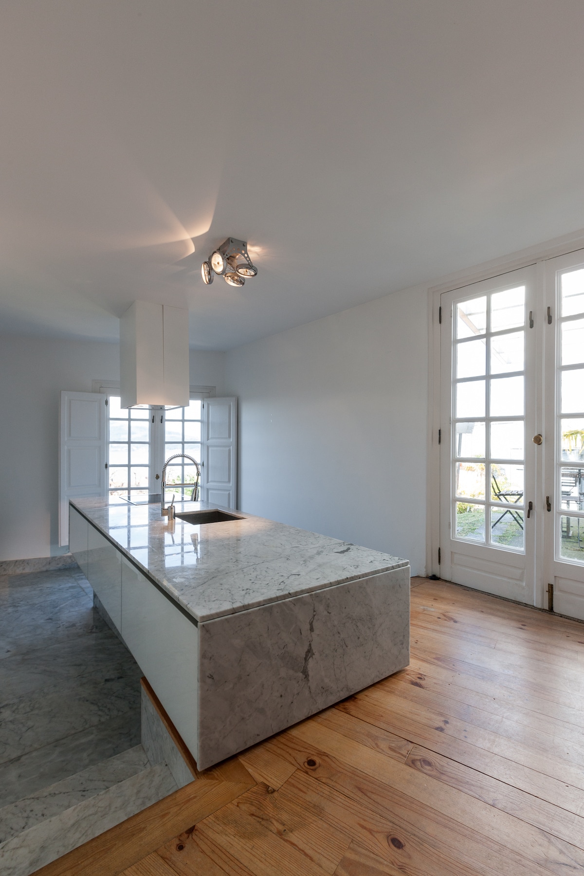 A modern kitchen features a central island with a marble countertop. Natural light floods the space through multiple glass doors that open to the outside, complementing the warm wooden flooring and creating a bright and airy atmosphere.