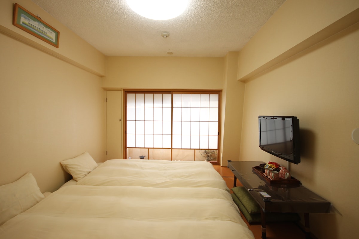 A bright room features two spacious beds placed side by side, adorned with neutral-colored bedding. Natural light filters through a traditional shoji window, creating a serene ambiance. A small television is positioned on a low table, adding to the cozy, functional layout of the space.