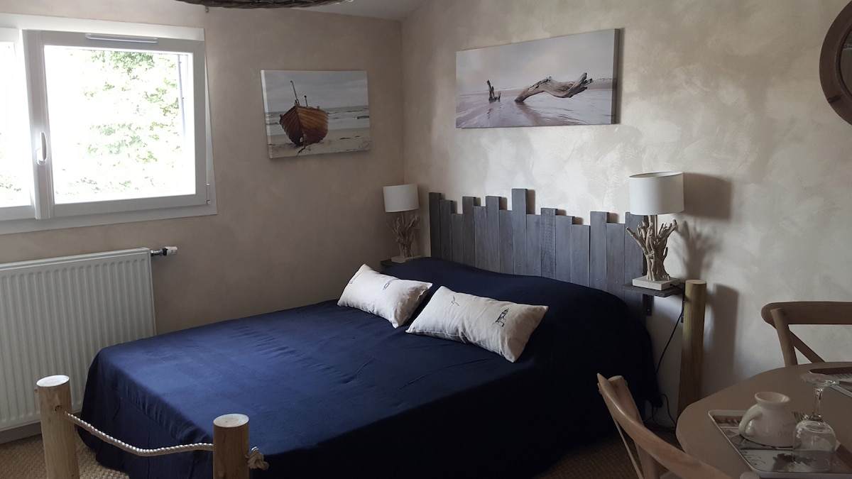 A calming bedroom features a spacious bed with navy blue bedding and decorative pillows. Two bedside lamps provide soft lighting. Artwork depicting coastal scenes is displayed above the bed, while a window allows natural light to illuminate the room. A small dining area is visible in the corner.