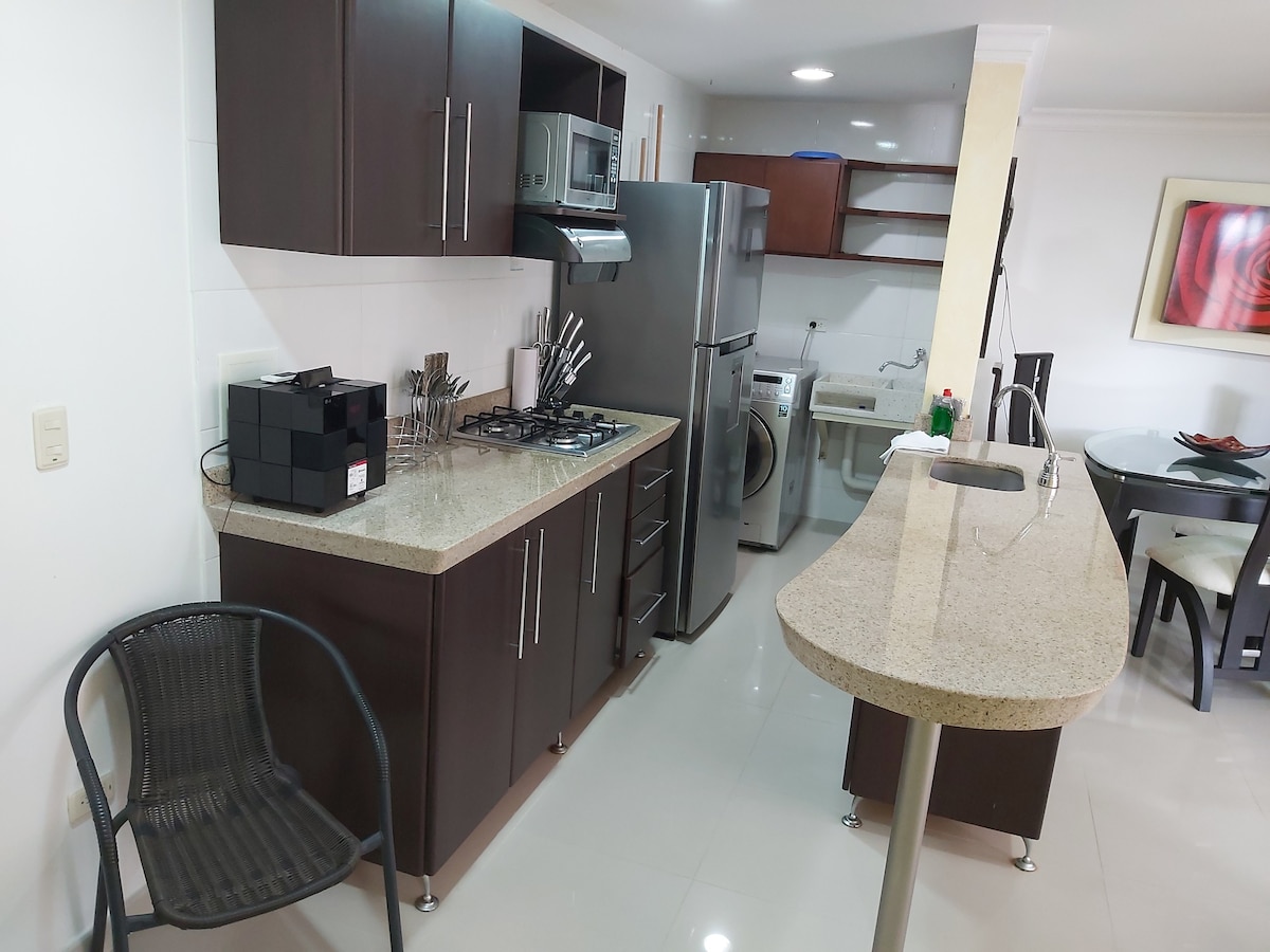 A modern kitchen is designed with dark cabinetry and a spacious countertop. Stainless steel appliances, including a refrigerator and gas stove, are prominently displayed. A dining area is visible, featuring a round table and chairs. Adequate lighting enhances the bright interior.