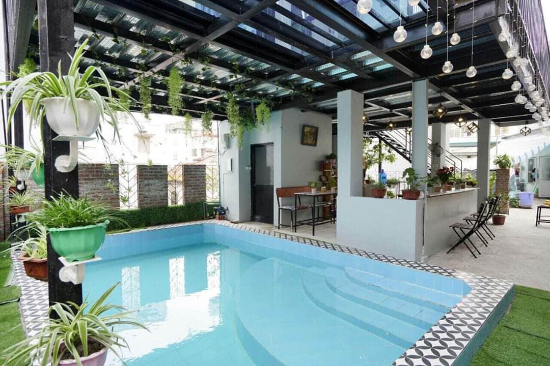 A shared outdoor area features a mini pool surrounded by greenery and potted plants. Comfortable seating and dining arrangements are visible under a covered structure, with string lights adding a soft illumination overhead. The space is designed for relaxation and socializing.