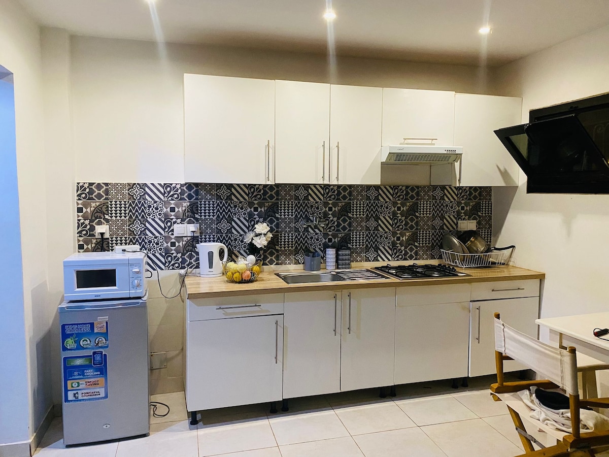 A modern kitchen area features sleek white cabinetry and a light wood countertop. Appliances include a microwave, kettle, and a gas stove with an extractor hood. A small refrigerator is positioned nearby, accompanied by a bowl of fruit and a patterned backsplash that adds visual interest.