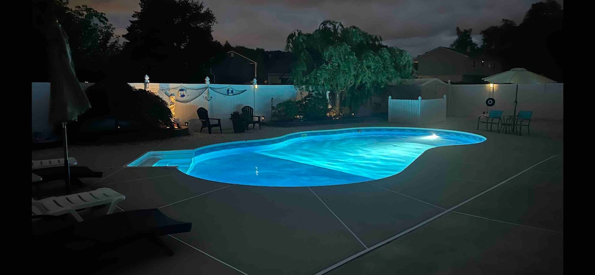 A private swimming pool is illuminated in soft blue light, creating a serene ambiance. Comfortable lounge chairs are positioned nearby, and lush greenery is visible around the pool area. The surrounding space offers fencing for privacy, enhancing the peaceful environment.