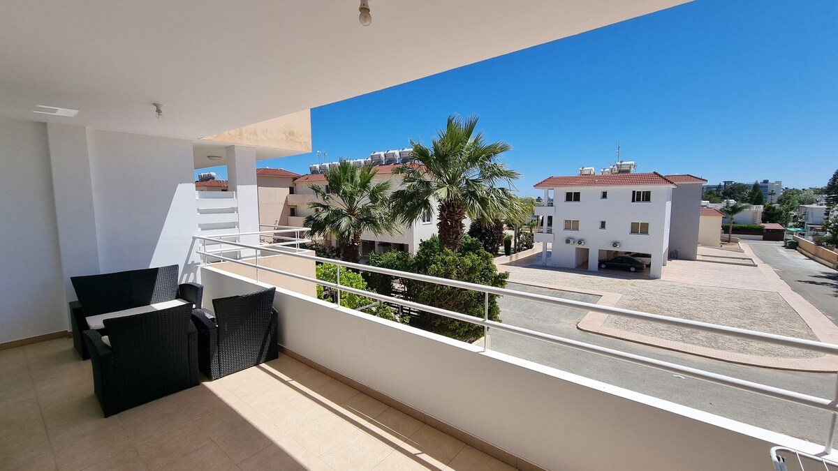 A balcony is visible, featuring a set of dark wicker chairs and a small table. The railing overlooks a well-maintained area with palms and buildings in the background, under a clear blue sky. The ground is paved, leading to additional structures.