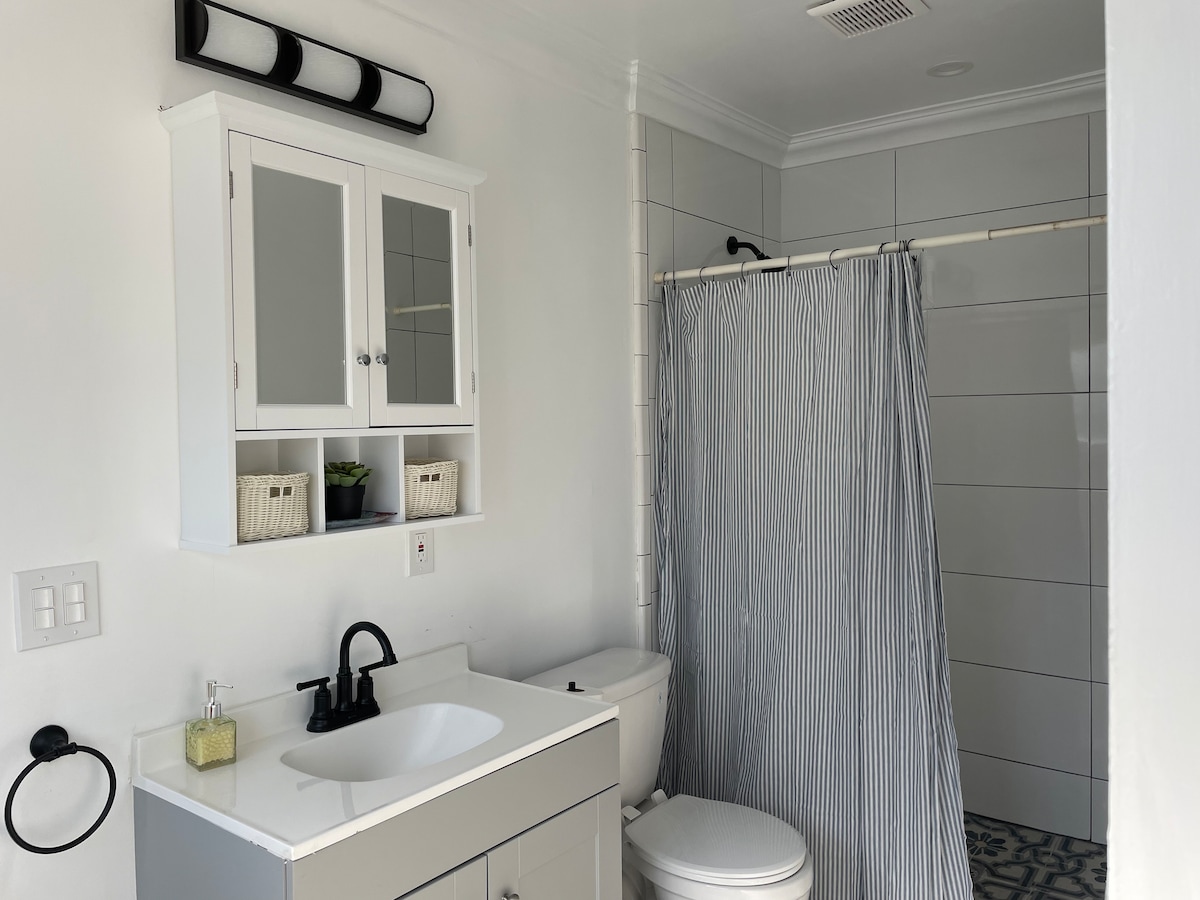 A recently renovated bathroom features a modern vanity with a sink and storage cabinet above. A striped shower curtain separates the shower area, while decorative baskets are displayed on the shelf. Neutral tones create a clean and tidy appearance.