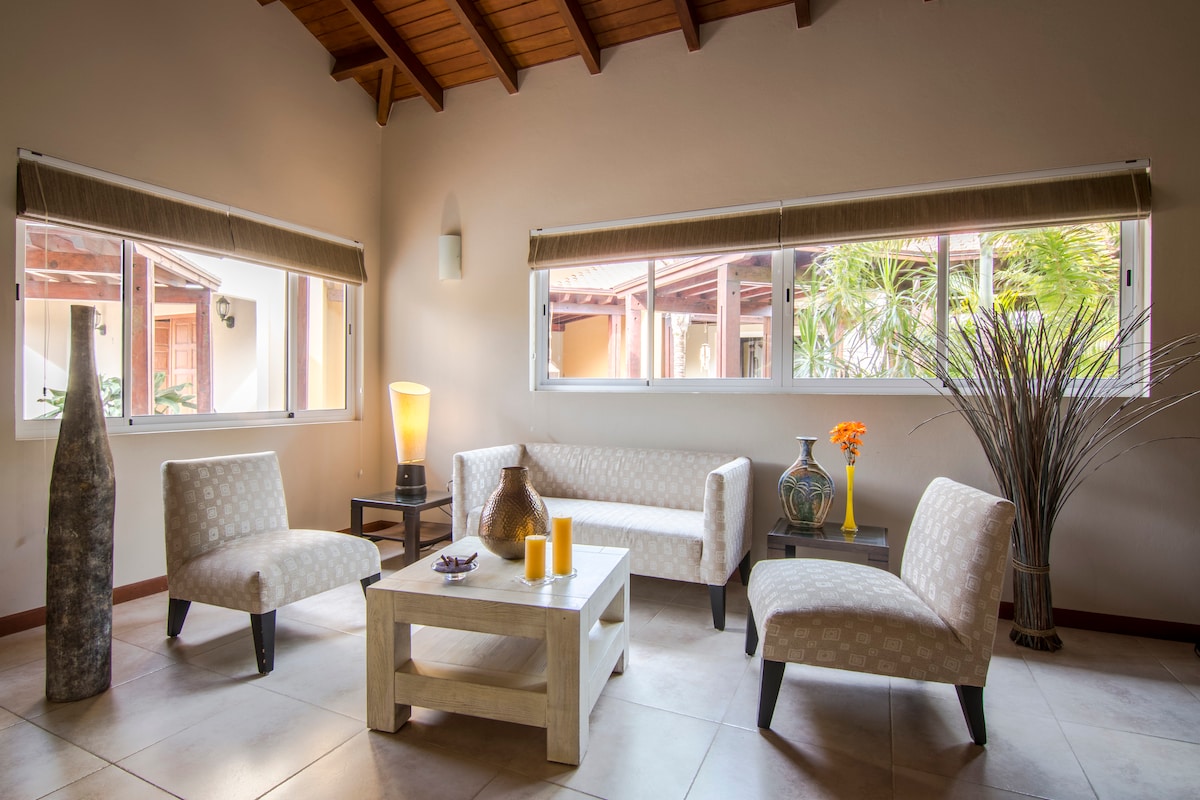 A welcoming living area is equipped with two patterned chairs, a sleek coffee table, and a sofa. Natural light filters through large windows adorned with patterned shades, highlighting decorative elements like a vase and a lamp. Tropical plants can be glimpsed outside, adding to the refreshing ambiance.