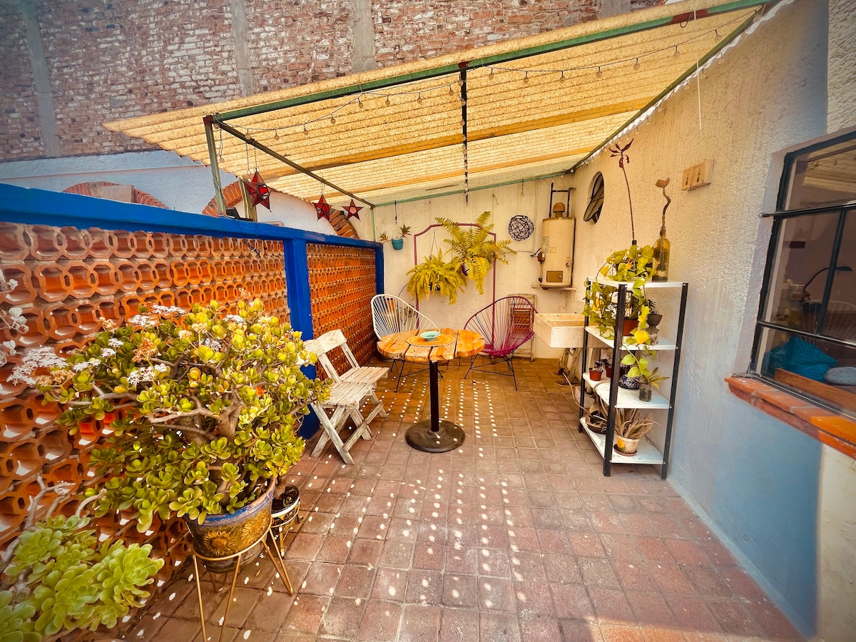 An outdoor patio area features a shaded seating arrangement with a round table and two chairs. Potted plants and decorative elements enhance the space, while sunlight filters through a canopy, creating a serene atmosphere. A brick wall adds texture to the backdrop.