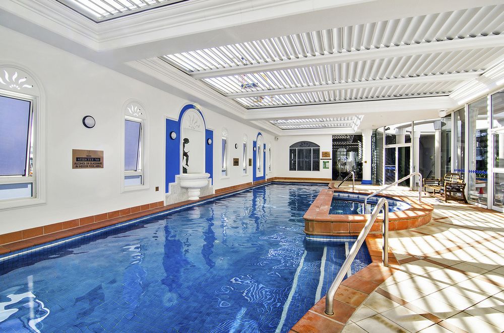 An indoor heated pool area is seen, featuring a large swimming pool with a clear blue surface. Natural light filters through skylights, illuminating the space. A hot tub is situated adjacent to the pool, with a handrail for safety. Decorative elements enhance the overall aesthetic.