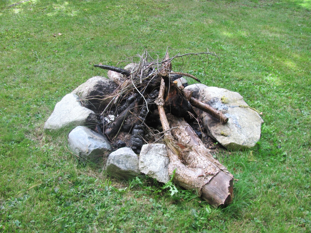 A campfire pit is surrounded by large, flat stones, featuring a gathering of logs and branches placed within. The grassy yard offers ample open space, framing the pit, inviting outdoor gatherings.