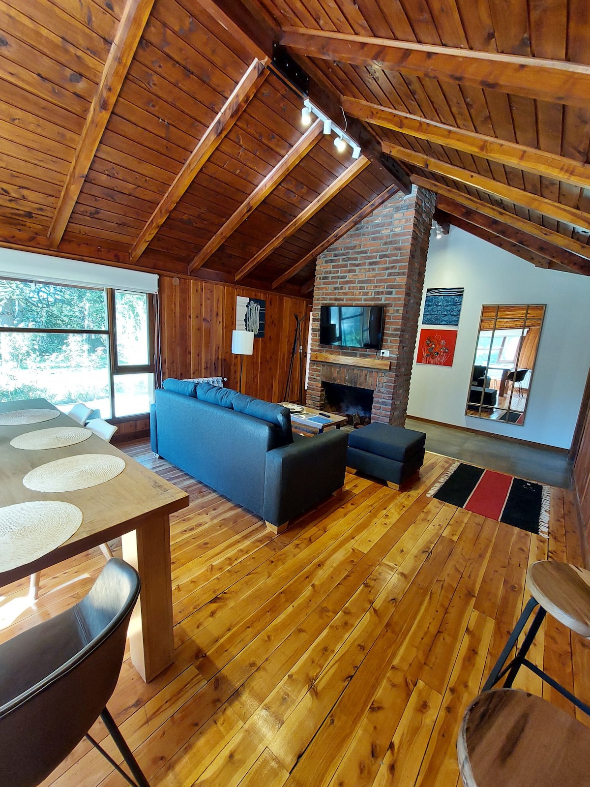 A spacious living area showcases wooden beams and a warm wood floor. A grey sofa is positioned in front of a brick fireplace, with a Smart TV mounted nearby. A dining table is set with placemats, while large windows allow natural light to fill the room.