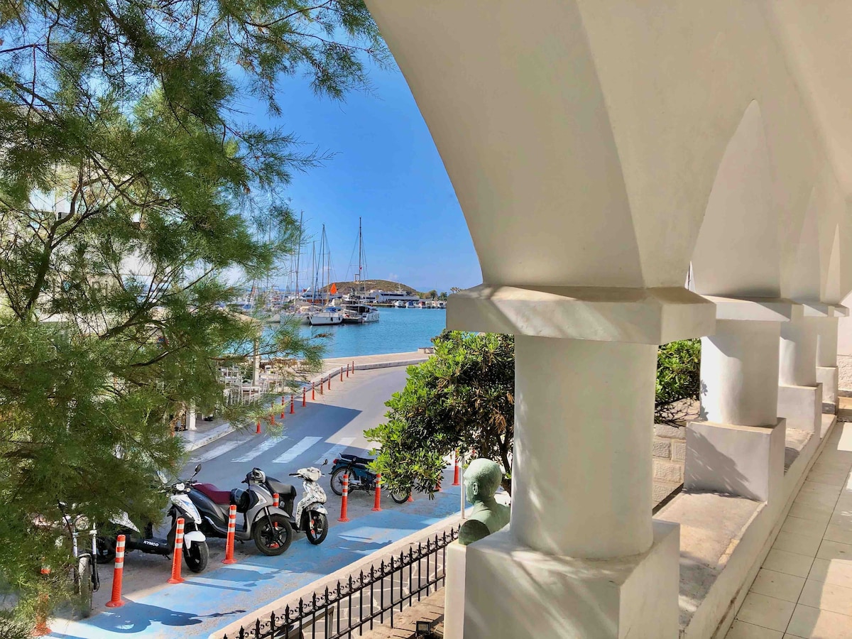 A view from an arched balcony showcases a vibrant scene of parked motorcycles and the marina in the distance. Boats are visible on the water, framed by greenery and supported by the architectural columns of the building.