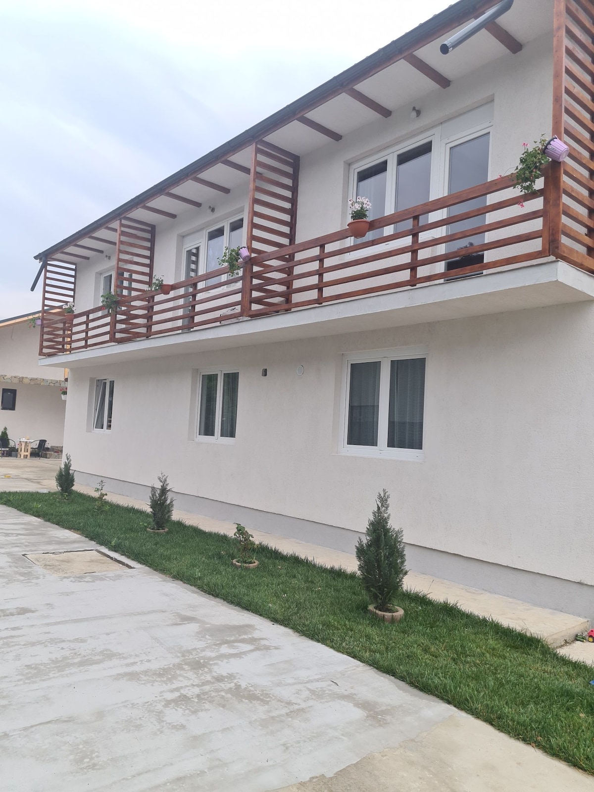 The exterior of a two-story building is showcased, featuring white walls and a wooden balcony adorned with potted plants. A well-maintained lawn with small shrubs lines the pathway, while several windows allow natural light to enter the space.