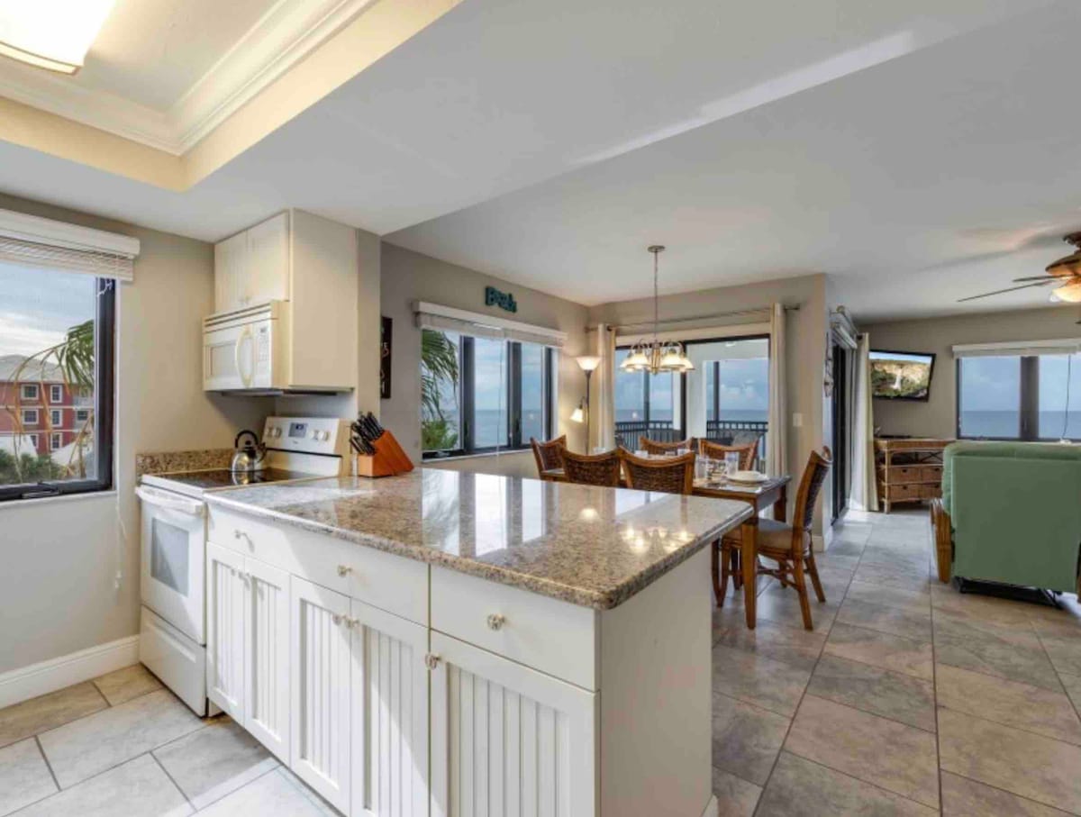 A bright kitchen is visible, featuring a spacious island with a granite countertop and white cabinetry. Natural light enters through the windows, revealing a dining area with a wooden table. Ocean views are present beyond the patio area, offering a serene backdrop.