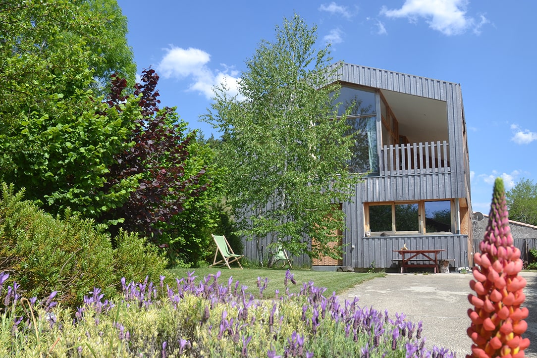 A modern wooden and glass structure is framed by vibrant greenery and colorful flowers. A spacious terrace is visible, complemented by a seating area in the garden, offering a serene and inviting setting.