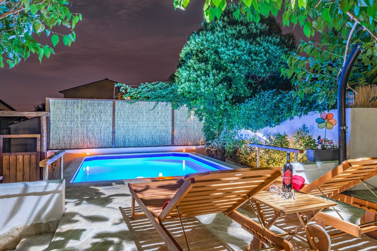 A small private swimming pool is surrounded by a paved area and wooden lounge chairs. Warm lighting highlights the water's surface, while a green tree offers shade. Decorative plants enhance the tranquil garden atmosphere, creating a peaceful outdoor space.