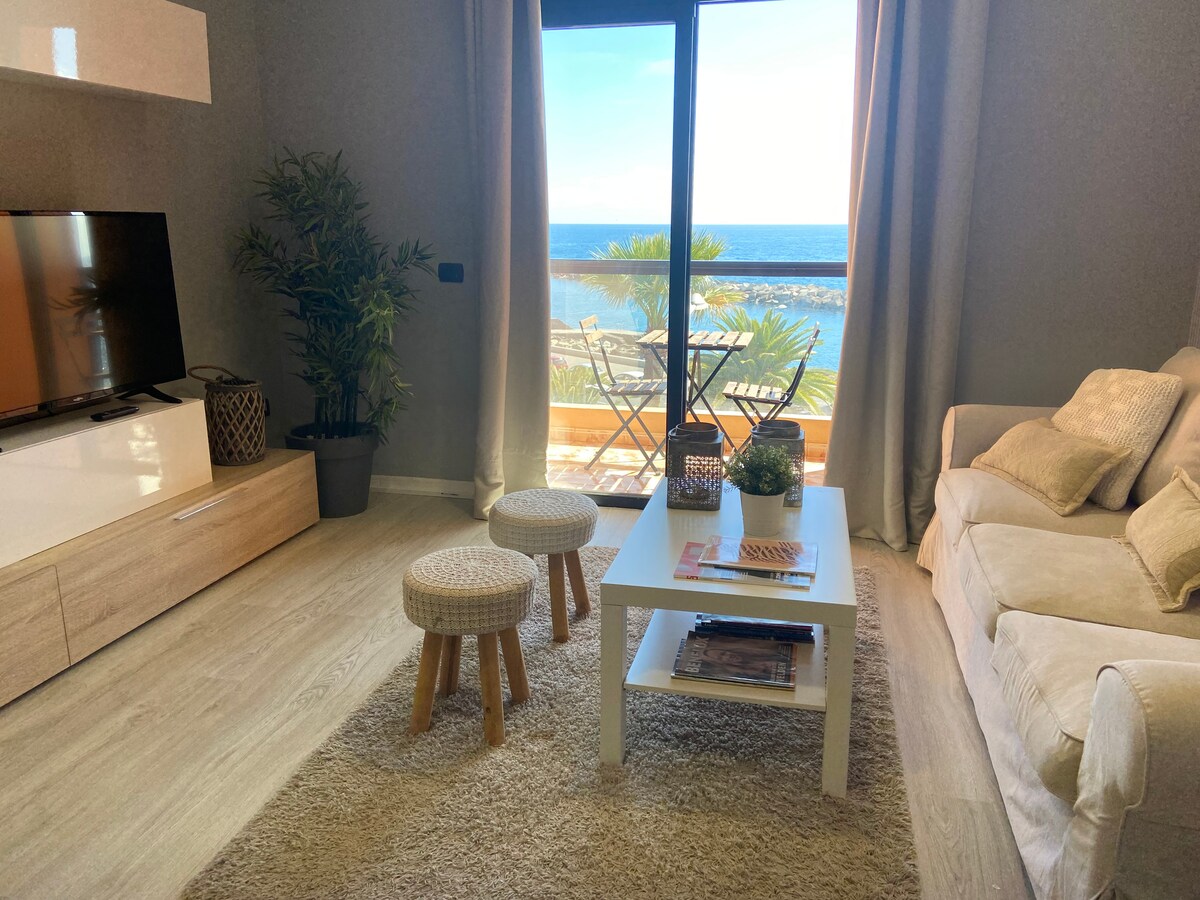 A light-filled living area features a comfortable sofa and a minimalist coffee table atop a soft rug. Large sliding doors open to a balcony with sea views, complemented by small tables and chairs outside. A television is positioned against a modern media unit.