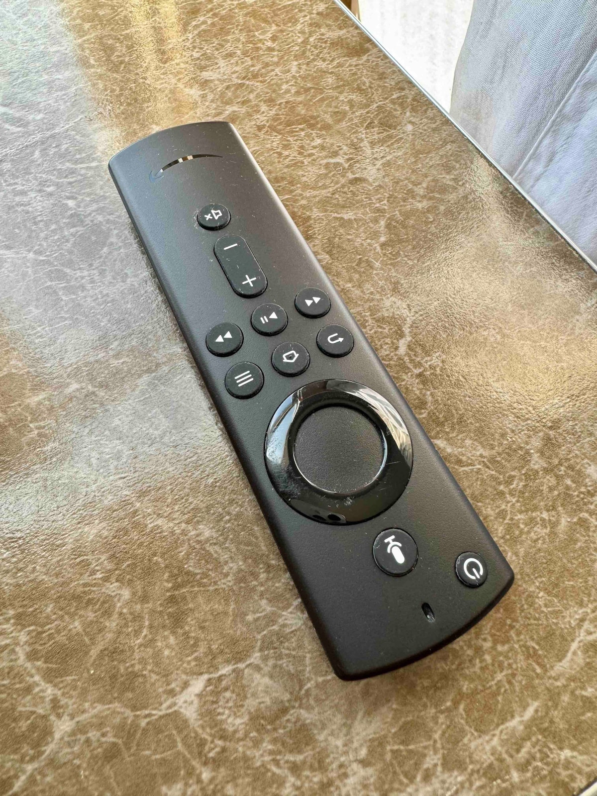 A black remote control is positioned on a textured surface, featuring various buttons including a circular navigation pad and control icons for easy operation. The background shows part of a window with soft, light-colored curtains allowing natural light to filter through.