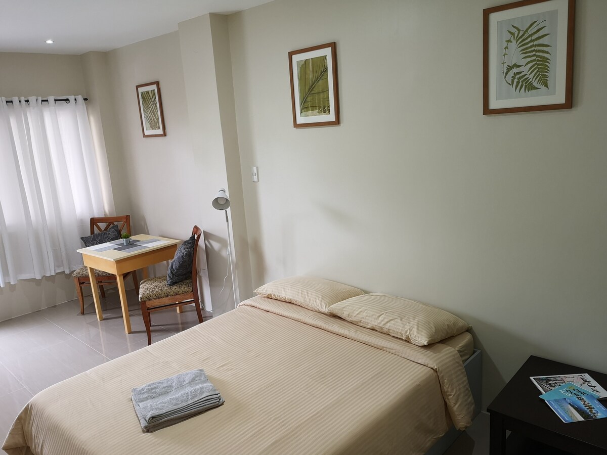 A cozy room features a spacious bed covered with a light-colored duvet. Natural light enters through sheer curtains, illuminating the neutral walls. A small dining table with two chairs is positioned near the window, accompanied by framed botanical artwork on the walls.