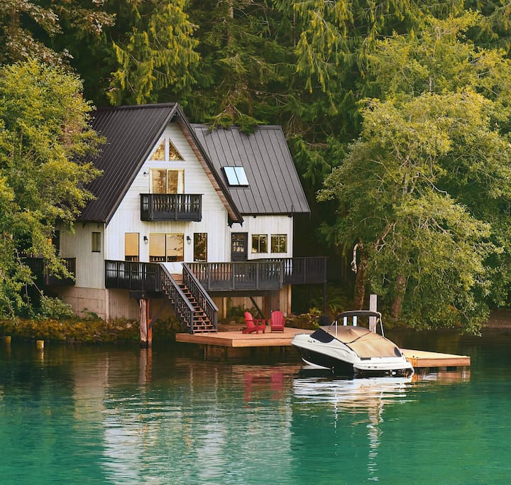 Une photo montre un bateau, couvert et amarré devant une maison de 2 étages située au bord d'un lac.