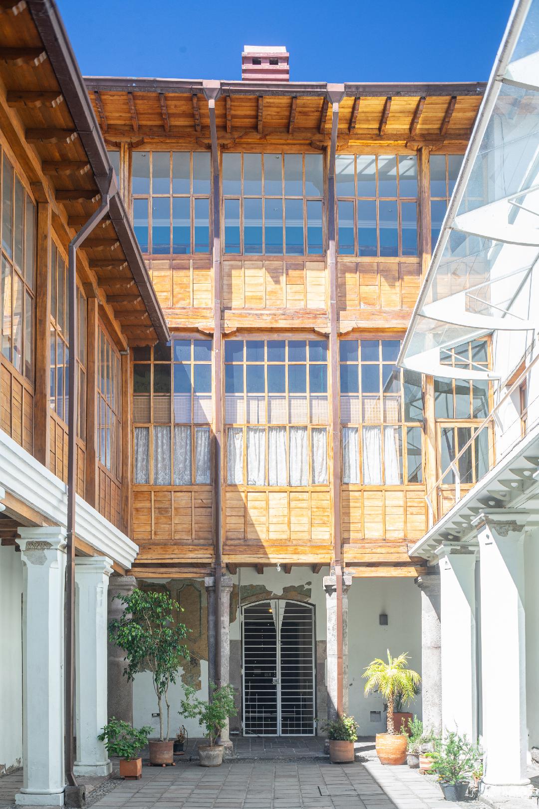 The image showcases a colonial-style structure with wooden beams and large windows, allowing ample natural light to fill the space. Potted plants are positioned at the base, contributing to the welcoming atmosphere of the central courtyard. The smooth stone flooring complements the architectural features.
