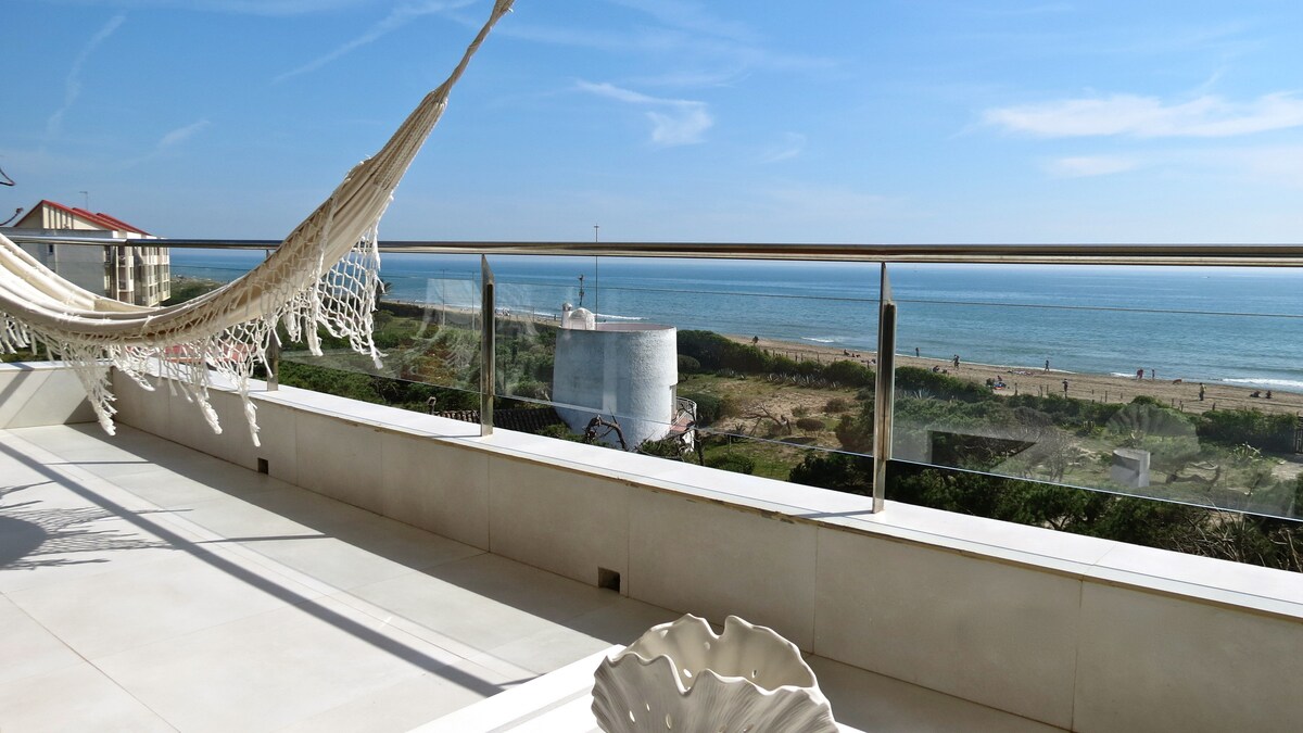 A spacious balcony offers unobstructed views of the beach and sea, with a hammock gently swaying against a backdrop of blue skies. The balcony is adorned with a decorative bowl, enhancing the serene coastal atmosphere.