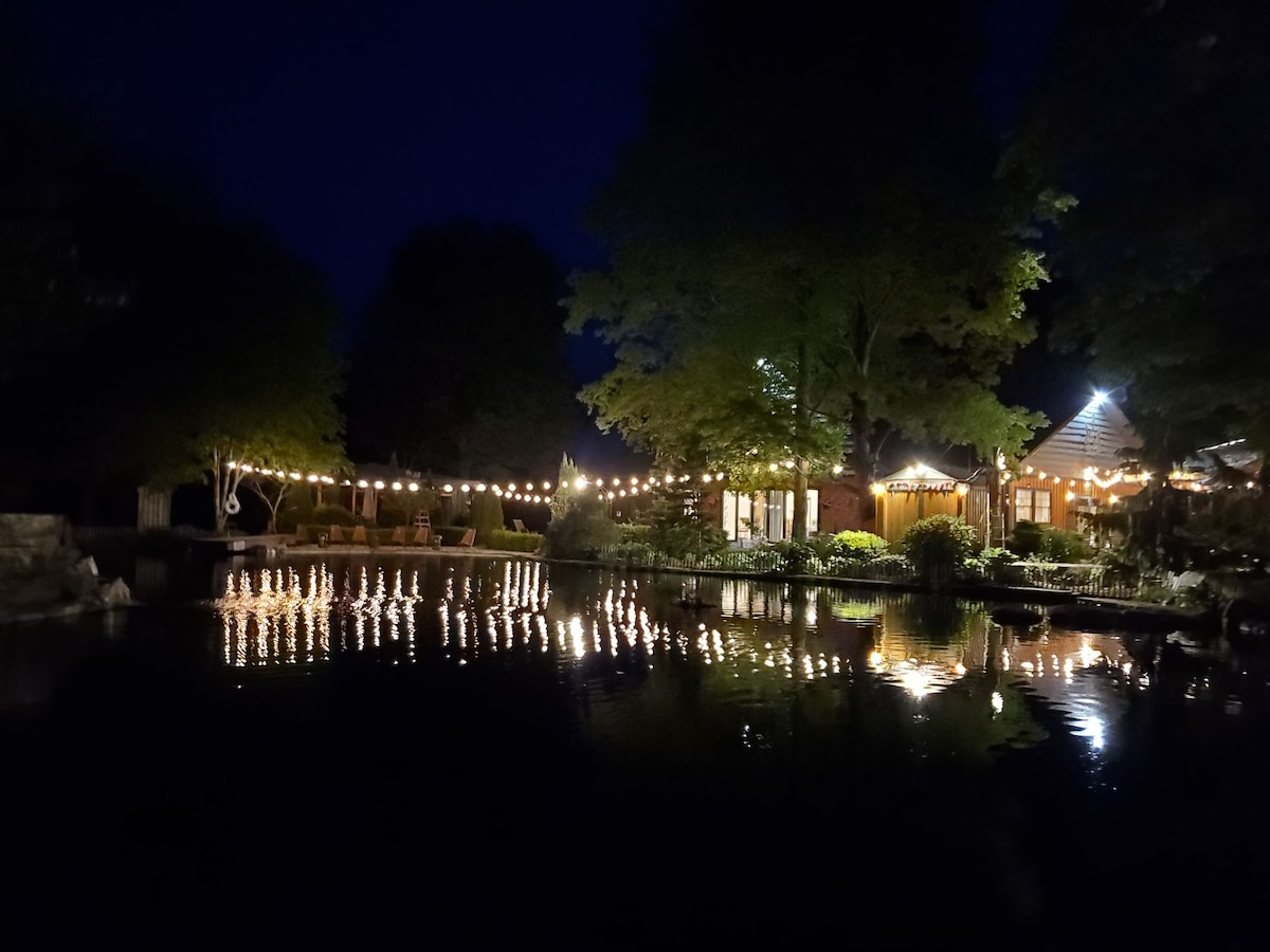 Softly illuminated pathways surround a tranquil pond at night, reflecting warm lights strung around the area. Lush trees border the scene, enhancing the serene atmosphere. The nearby structure glows gently, contributing to the peaceful ambiance of the outdoor space.