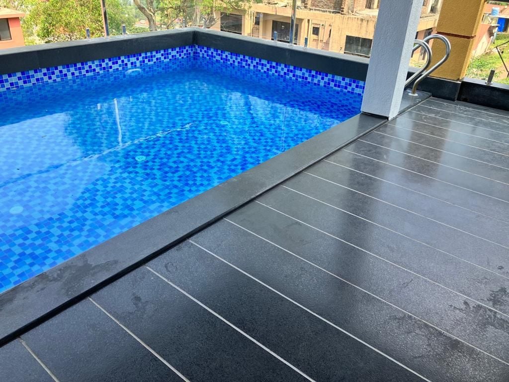 A private pool is visible, featuring vibrant blue water and a mosaic tile border. The surrounding area consists of sleek dark stone flooring, enhancing the modern aesthetic. Sunlight reflects off the water's surface, creating a serene setting.