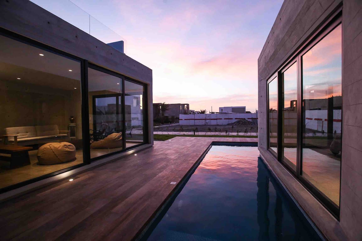 A private pool reflects the colors of the sunset, bordered by modern glass walls. The outdoor deck features a seamless transition to a spacious living area, complemented by natural light streaming through large windows.